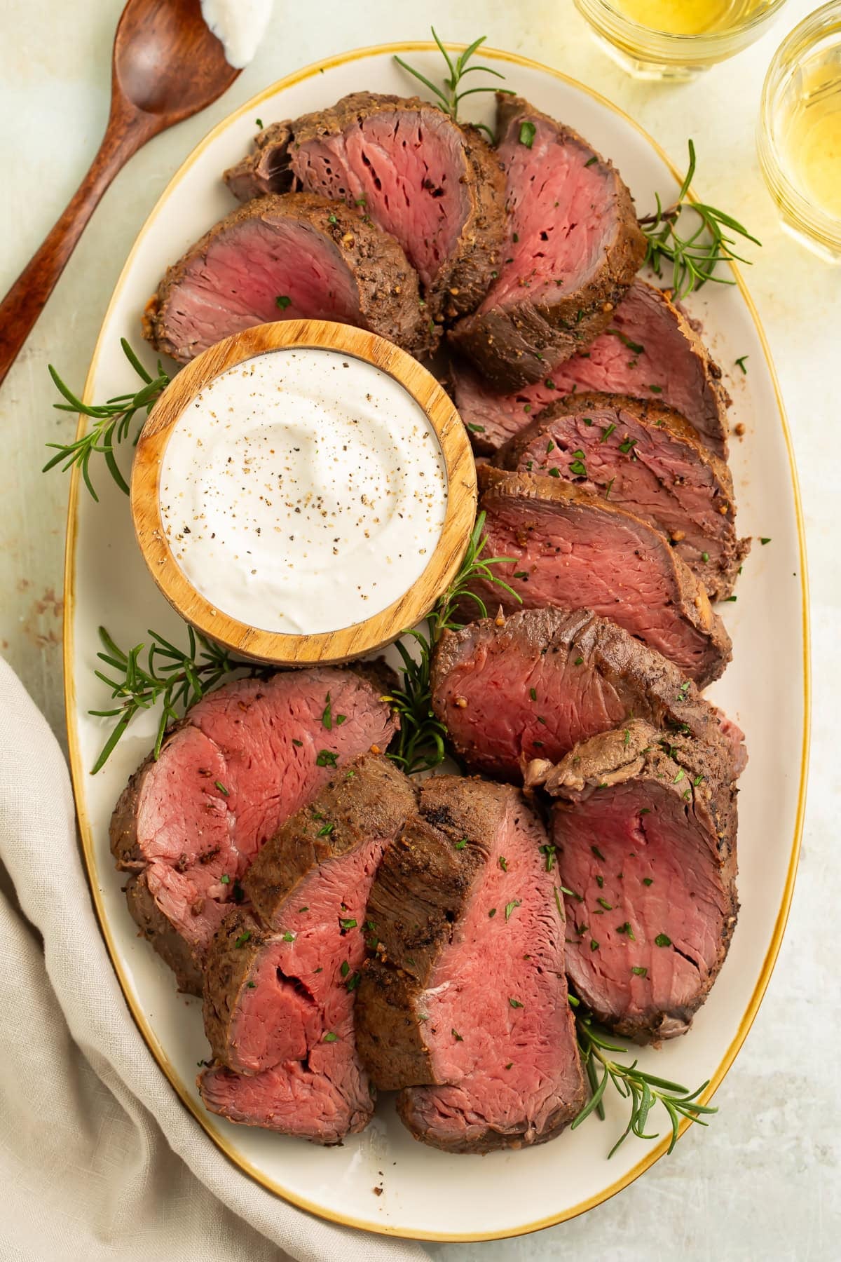 Overhead view of sliced smoked beef tenderloin arranged around small wooden bowl of horseradish sauce on white serving platter
