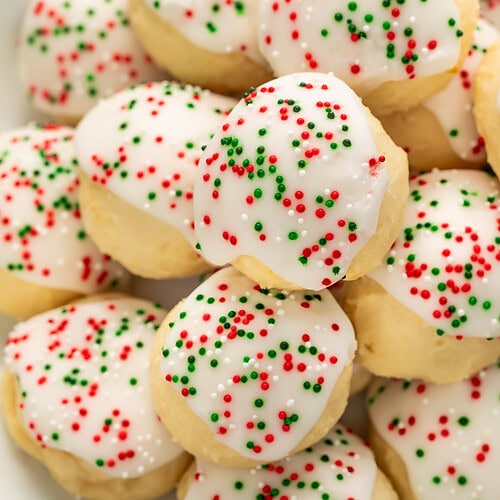 Pile of soft Italian Christmas cookies with white almond glaze and red and green sprinkles on white plate