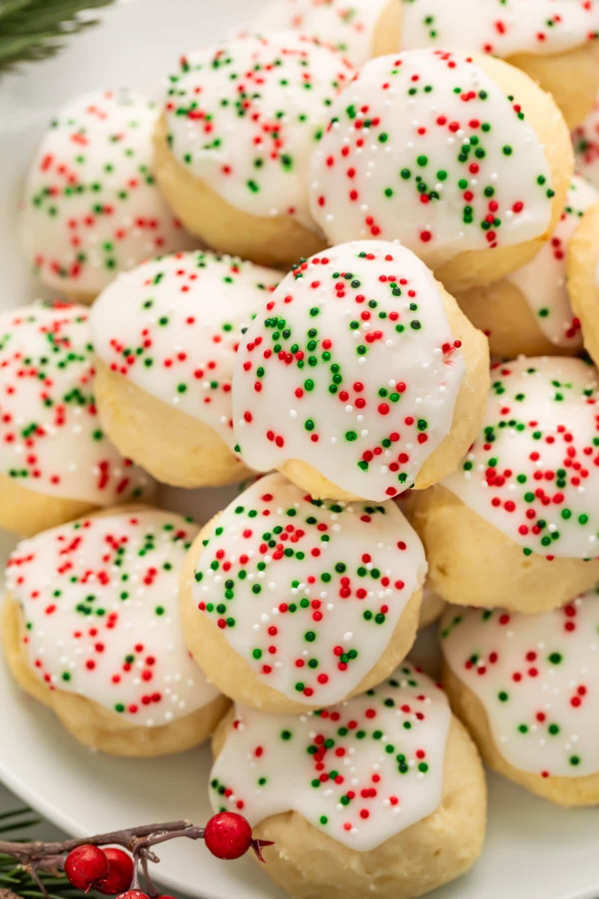 Pile of soft Italian Christmas cookies with white almond glaze and red and green sprinkles on white plate