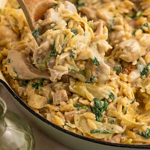 Wooden spoon lifting creamy chicken orzo pasta with spinach and artichokes from white skillet