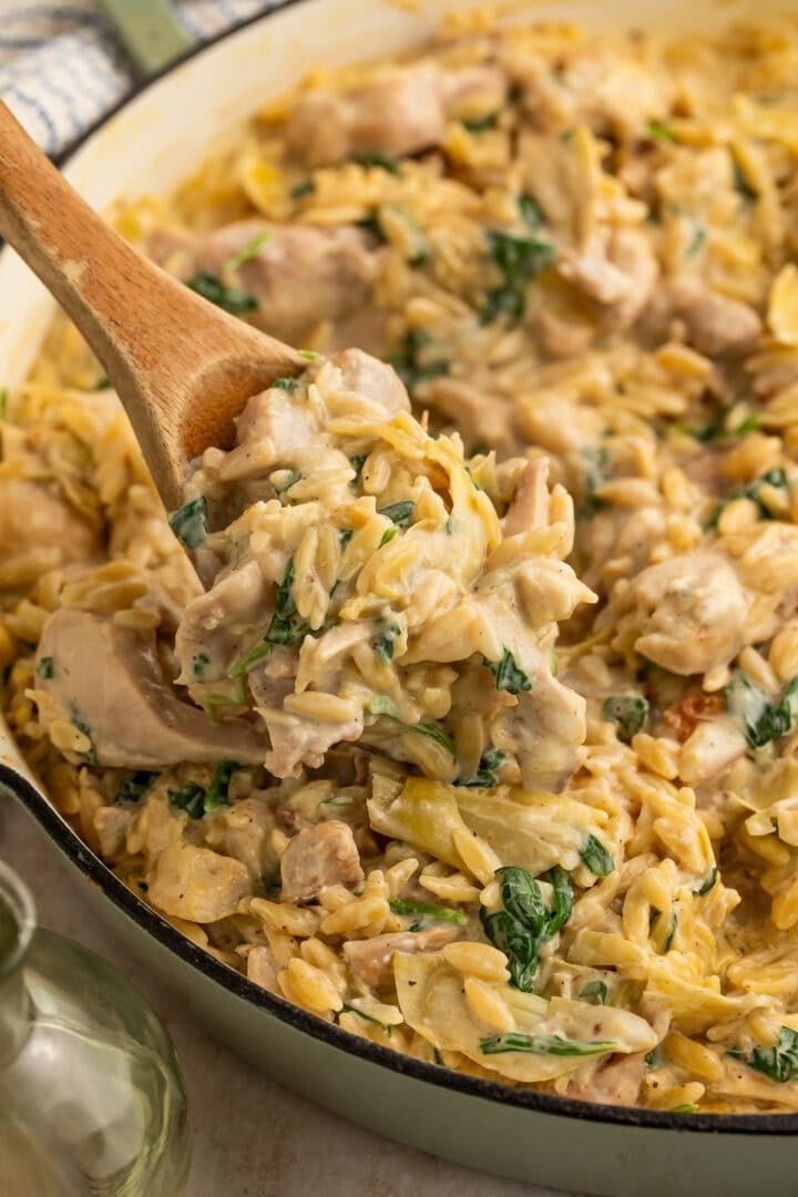 Wooden spoon lifting creamy chicken orzo pasta with spinach and artichokes from white skillet