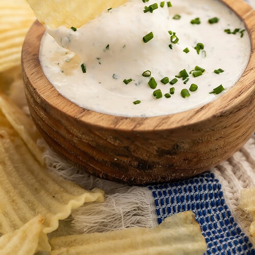 Hand holding ridged potato chip dipping into creamy herb dip in wooden bowl with fresh chives.
