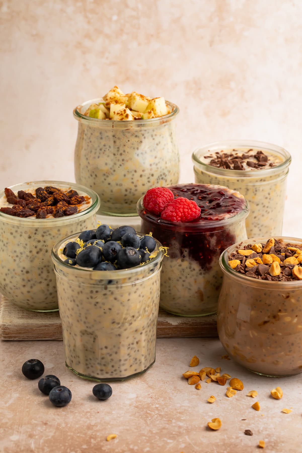 Six glass jars of overnight oats with different toppings including blueberries, raspberries, apples, chocolate, and raisins