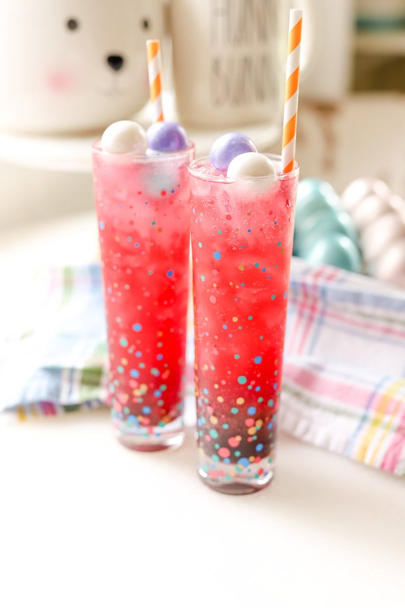 Two tall glasses of pink Easter mocktail with pastel candy eggs and orange striped straws on a colorful plaid napkin