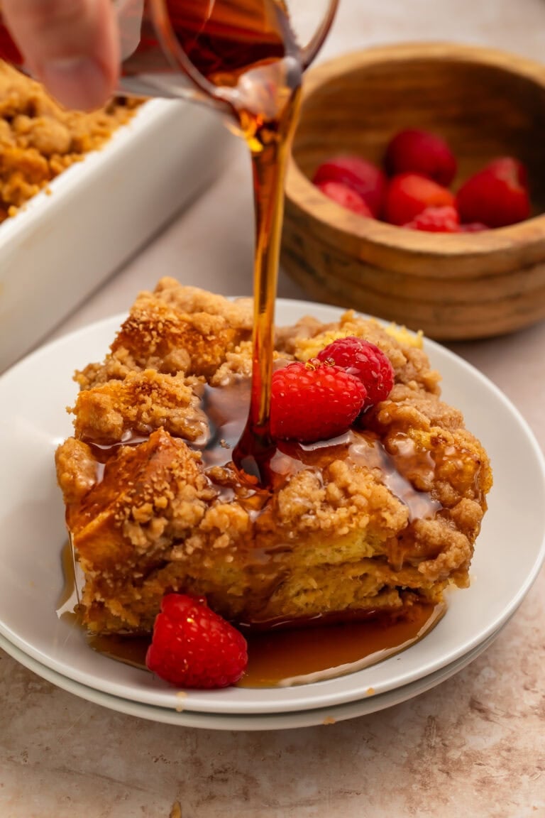 Maple syrup pouring over a slice of brioche French toast casserole with raspberries on a white plate