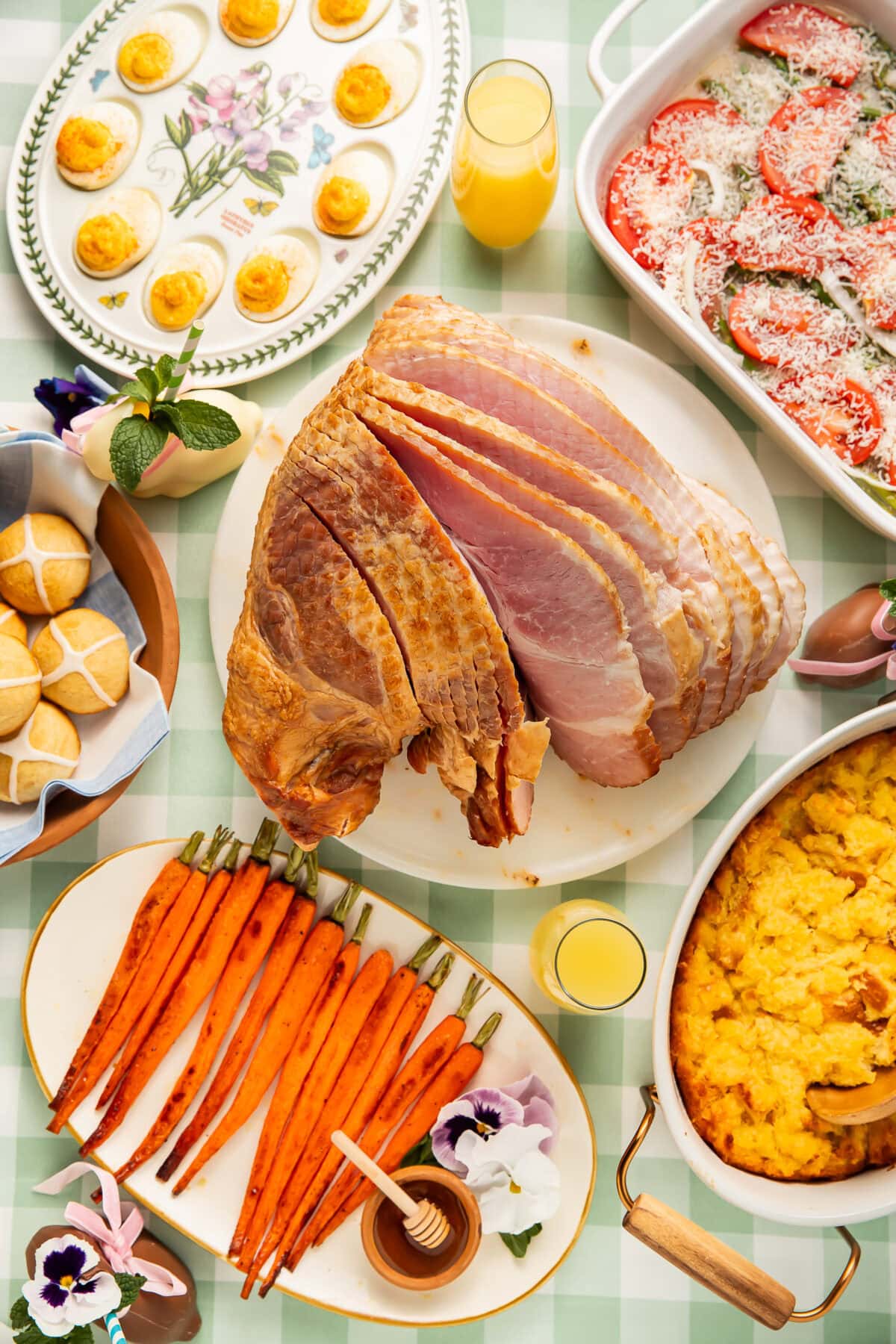 Overhead view of an Easter dinner spread with spiral ham, deviled eggs, roasted carrots, hot cross buns, and casserole dishes on a green gingham tablecloth