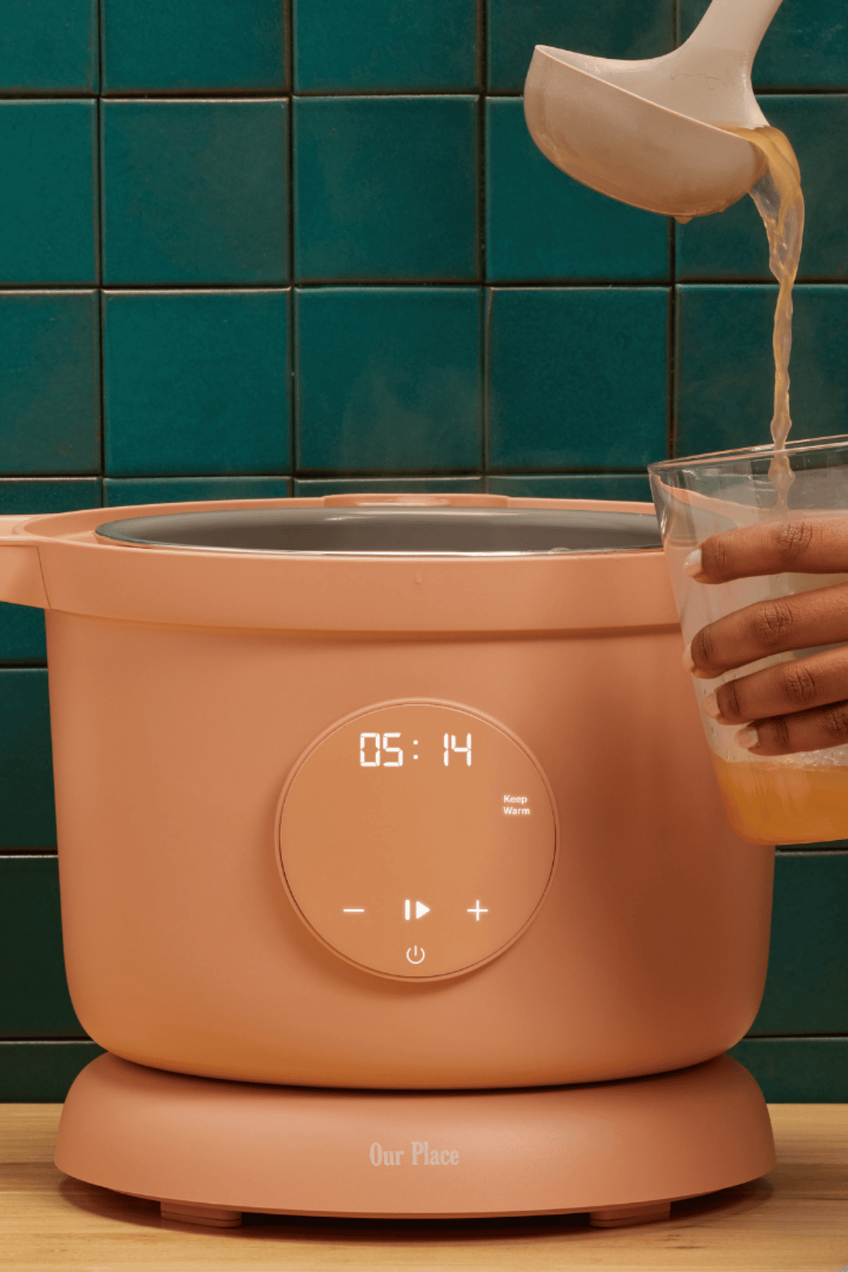 Liquid being poured into an open Our Place Dream Cooker against a teal tile wall