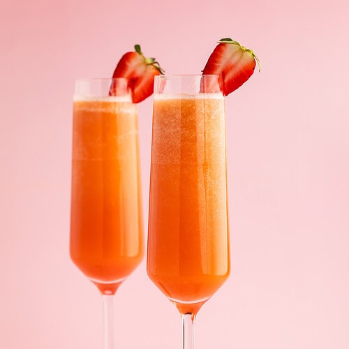 Two strawberry mimosas in champagne flutes garnished with halved strawberries on a pink background