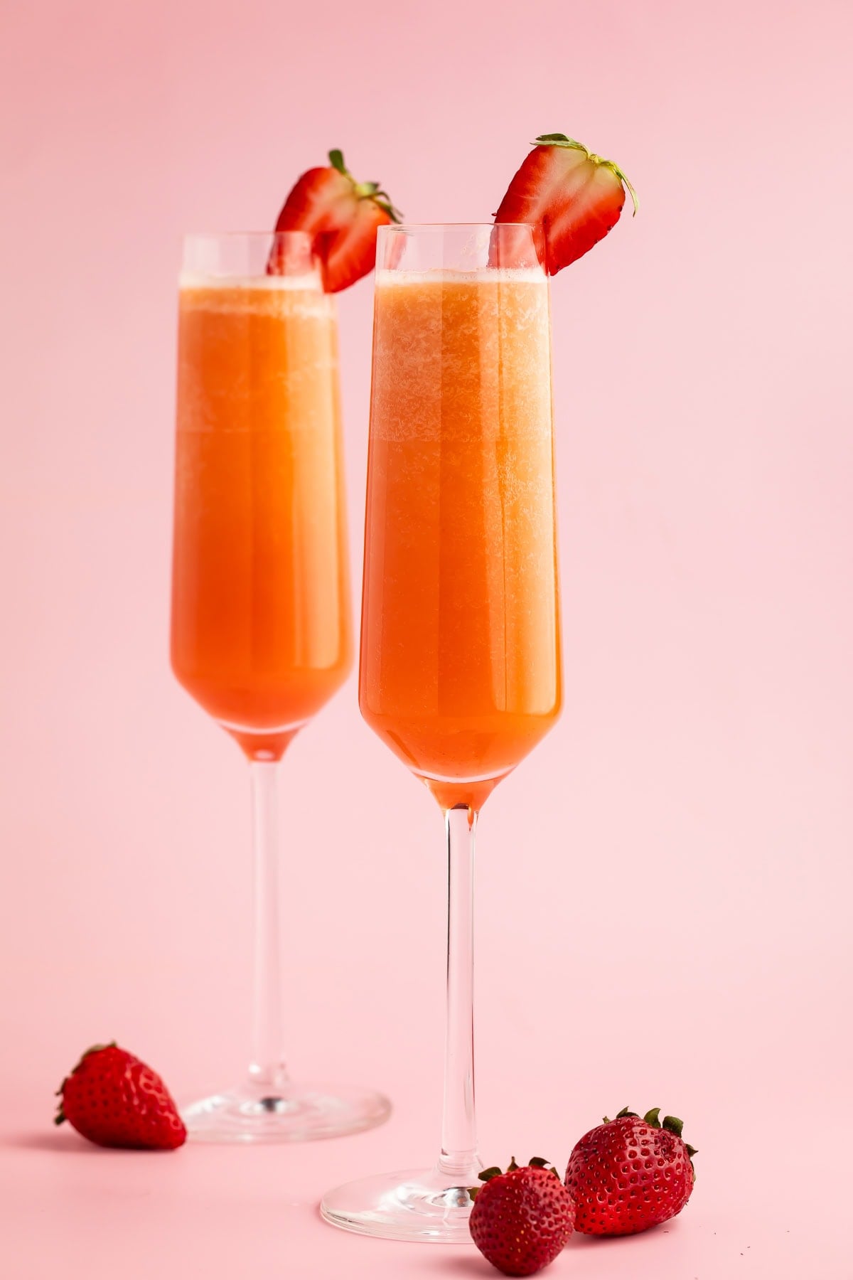 Two strawberry mimosas in champagne flutes garnished with halved strawberries on a pink background
