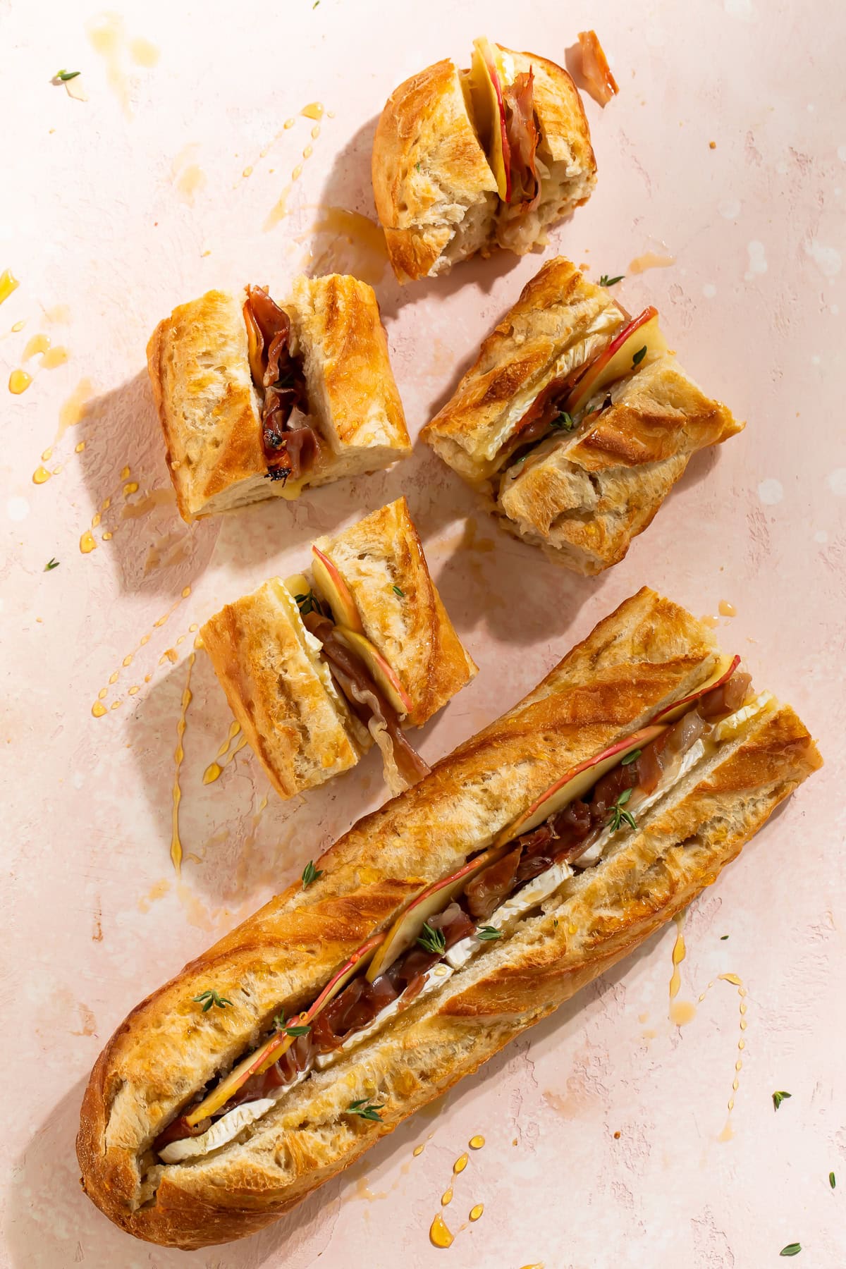 Overhead flat lay of a whole baked brie and prosciutto stuffed baguette with individual sliced portions on a pink surface