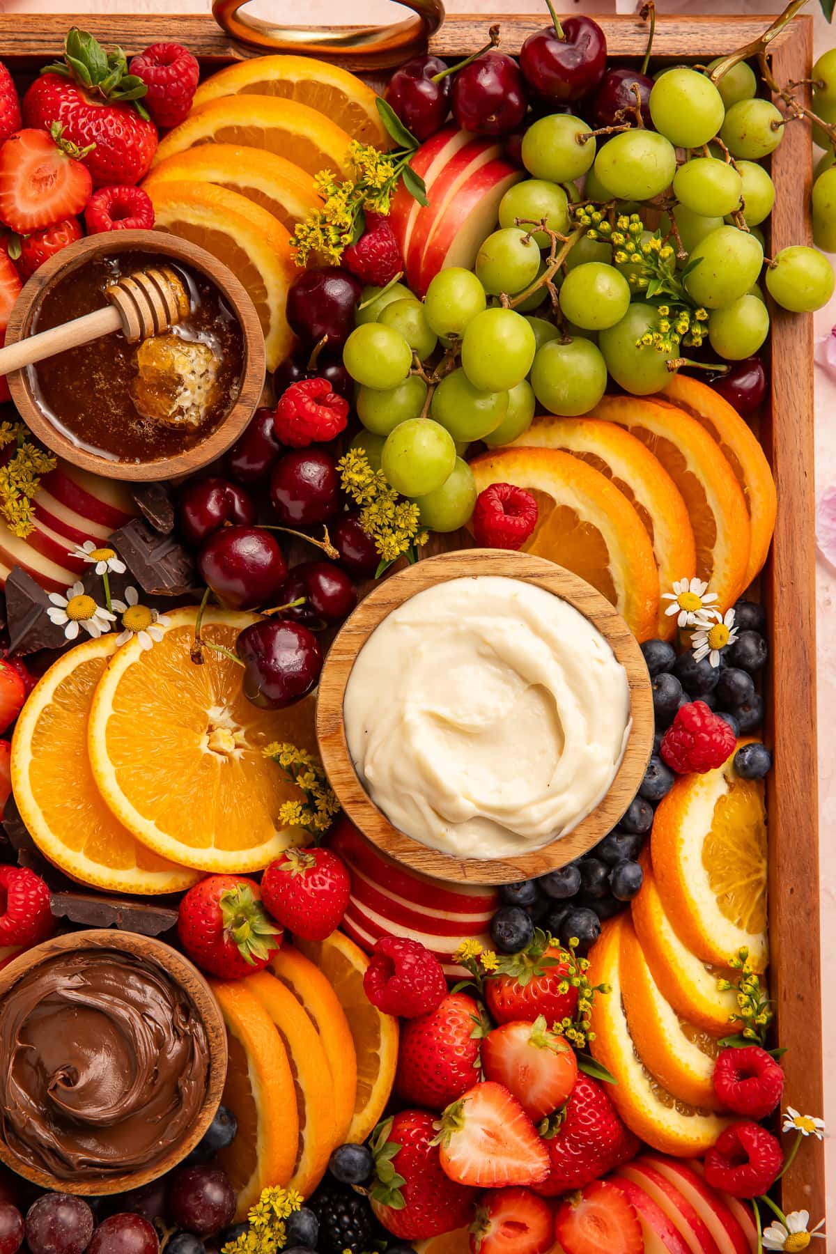 Overhead close-up of a wooden fruit board with orange slices, green grapes, cherries, berries, and dipping bowls