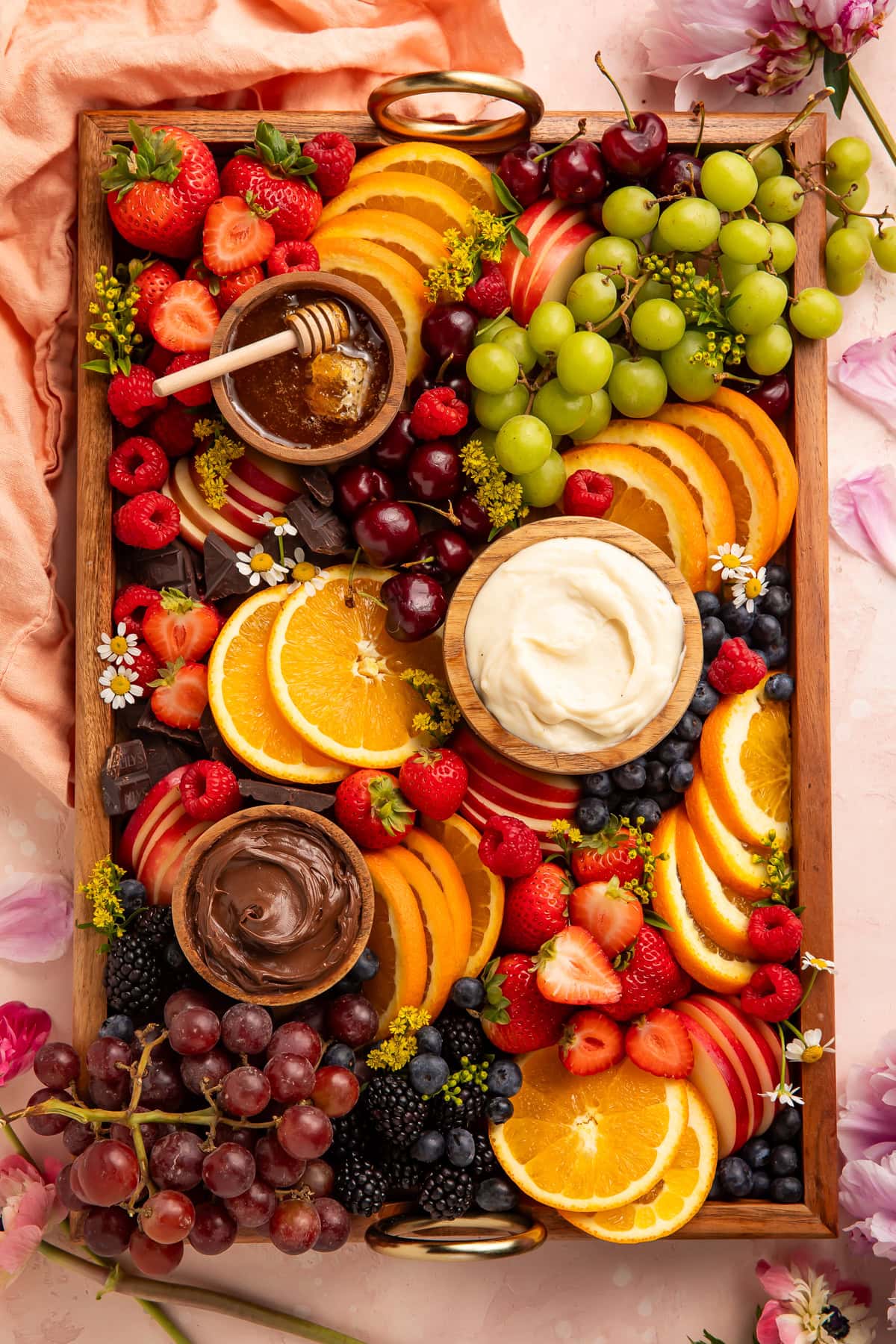 Full overhead view of a wooden tray fruit board with orange slices, mixed berries, grapes, and three dipping bowls surrounded by flowers