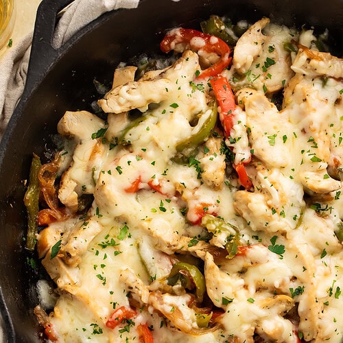 Overhead shot of chicken Philly cheesesteak skillet in cast iron pan with melted cheese and fresh parsley on linen