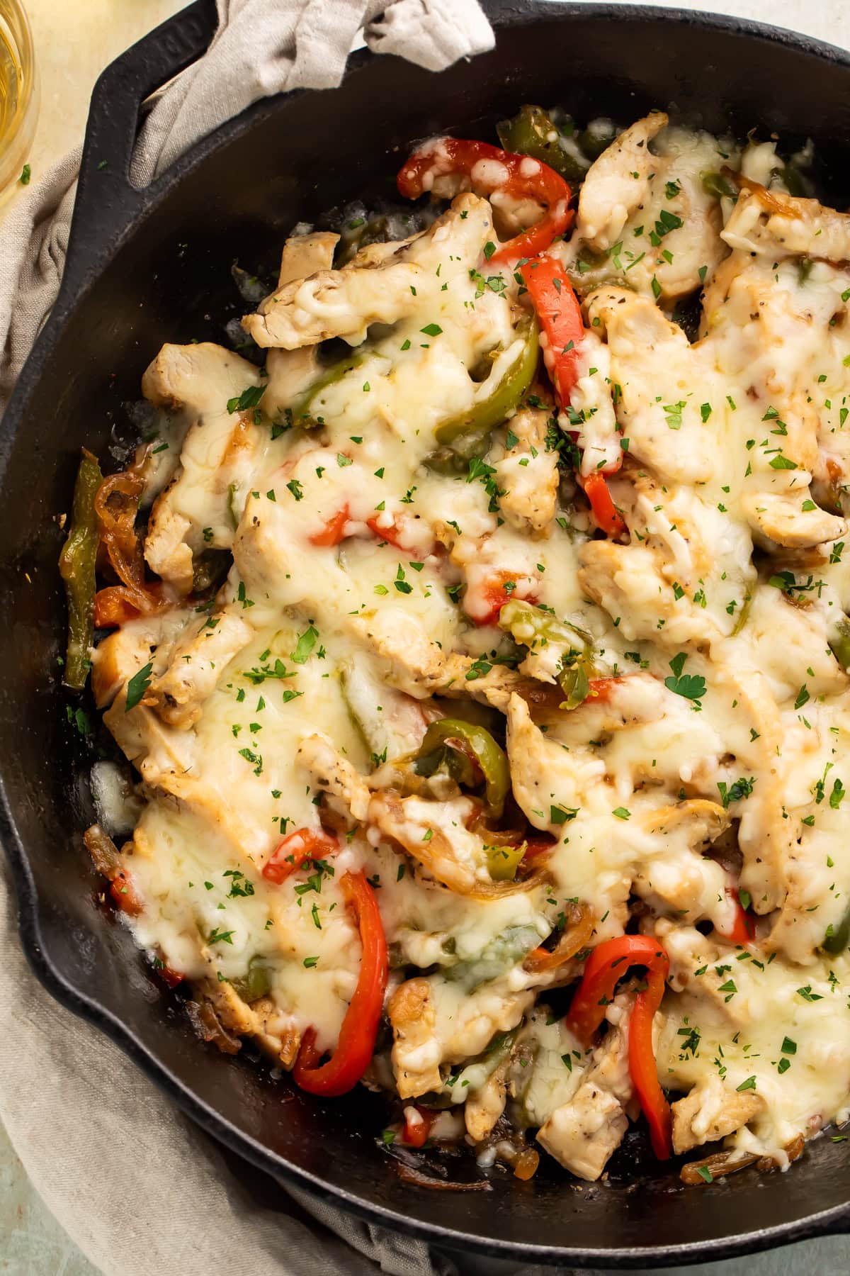 Overhead shot of chicken Philly cheesesteak skillet in cast iron pan with melted cheese and fresh parsley on linen