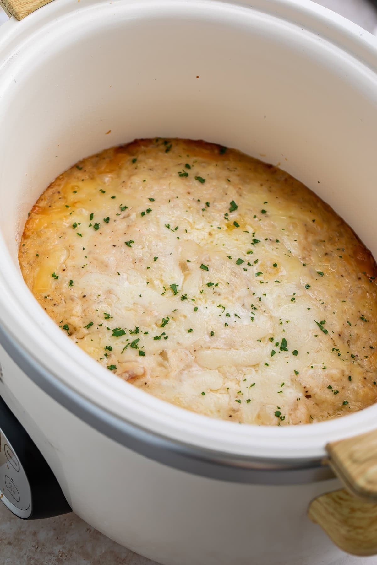 Crockpot scalloped potatoes with a golden melted cheese top in a white oval slow cooker, garnished with parsley