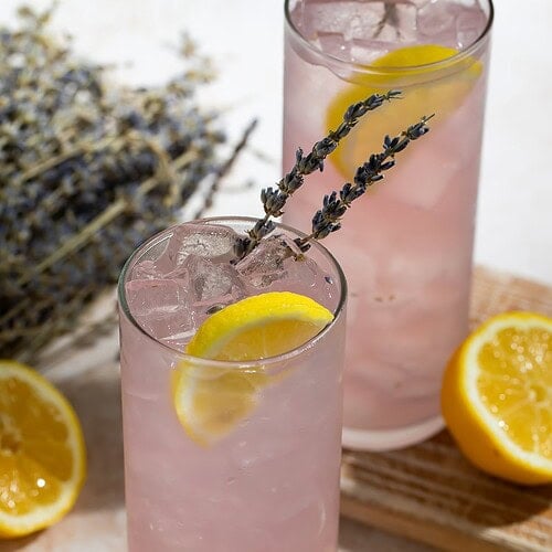 Two lavender lemonade cocktails in highball glasses garnished with lemon wedges and dried lavender sprigs