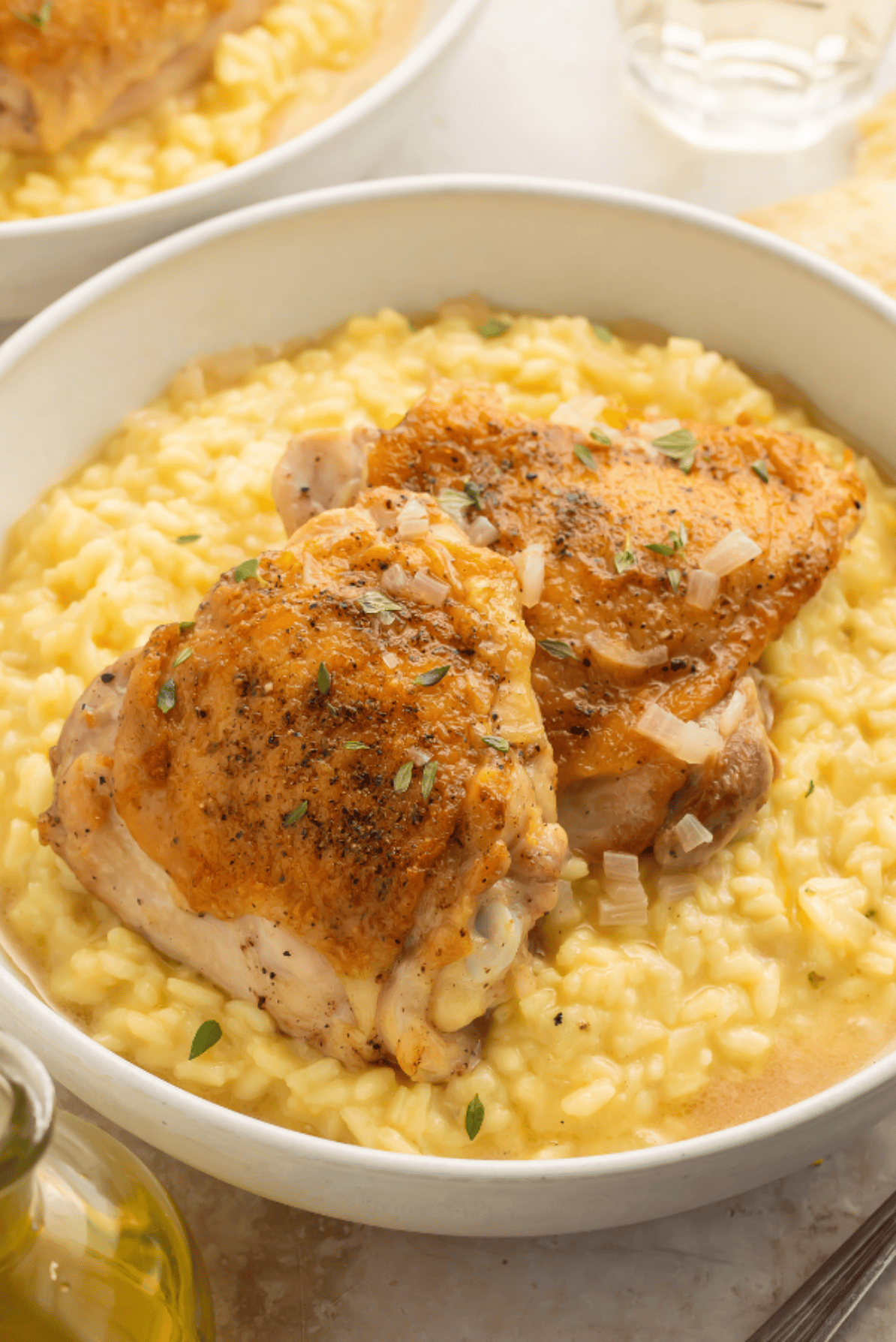 Two golden-brown chicken thighs served over creamy yellow risotto in a white bowl garnished with fresh thyme