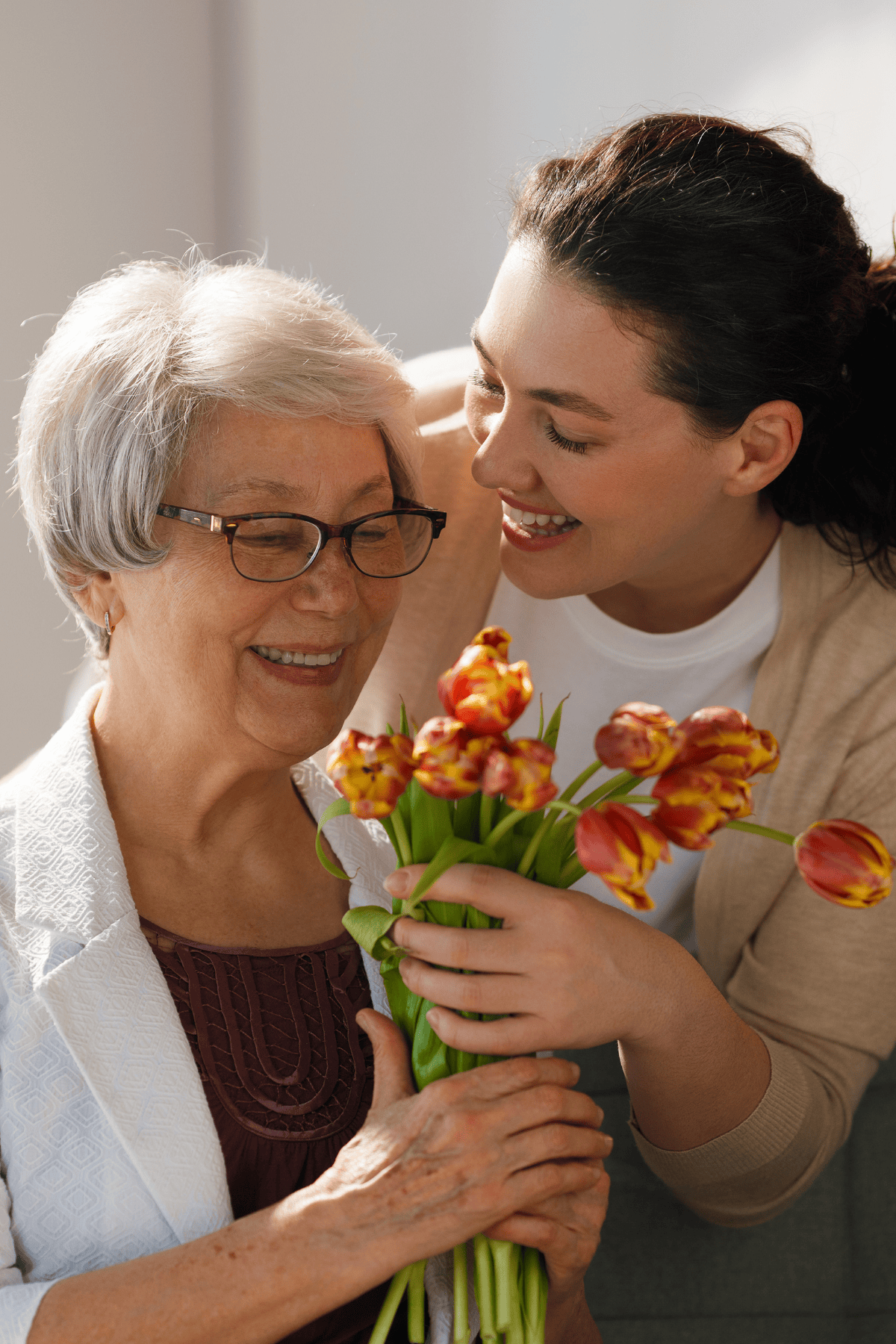An older woman with gray hair and glasses and a younger dark-haired woman smiling together holding red and yellow tulips