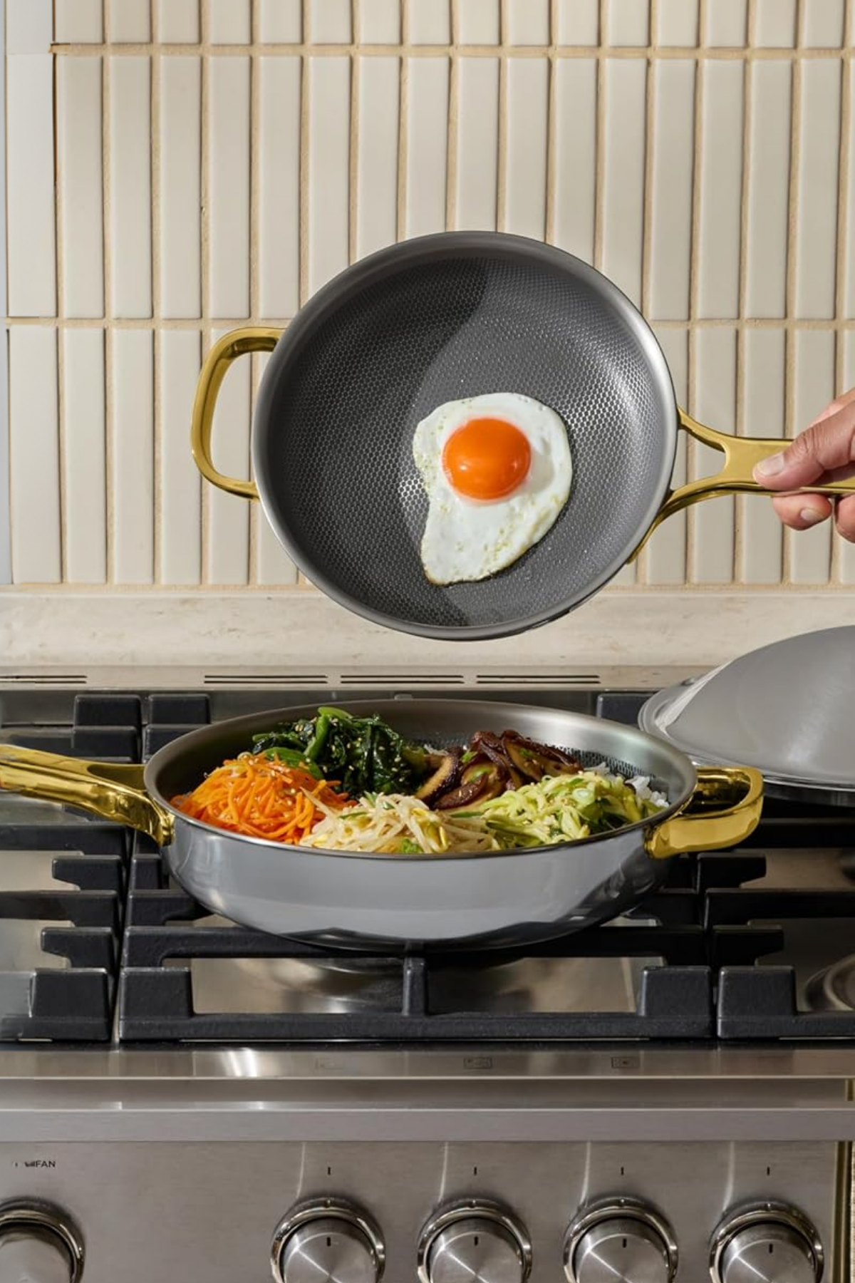 Two titanium pans with gold handles on a gas stovetop, one with a fried egg and one with bibimbap-style vegetables