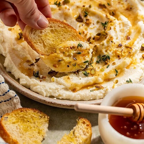 Overhead view of whipped ricotta dip in a shallow bowl, drizzled with honey and topped with chopped pistachios, fresh thyme, and red pepper flakes, with toasted baguette slices and lemon wedges alongside
