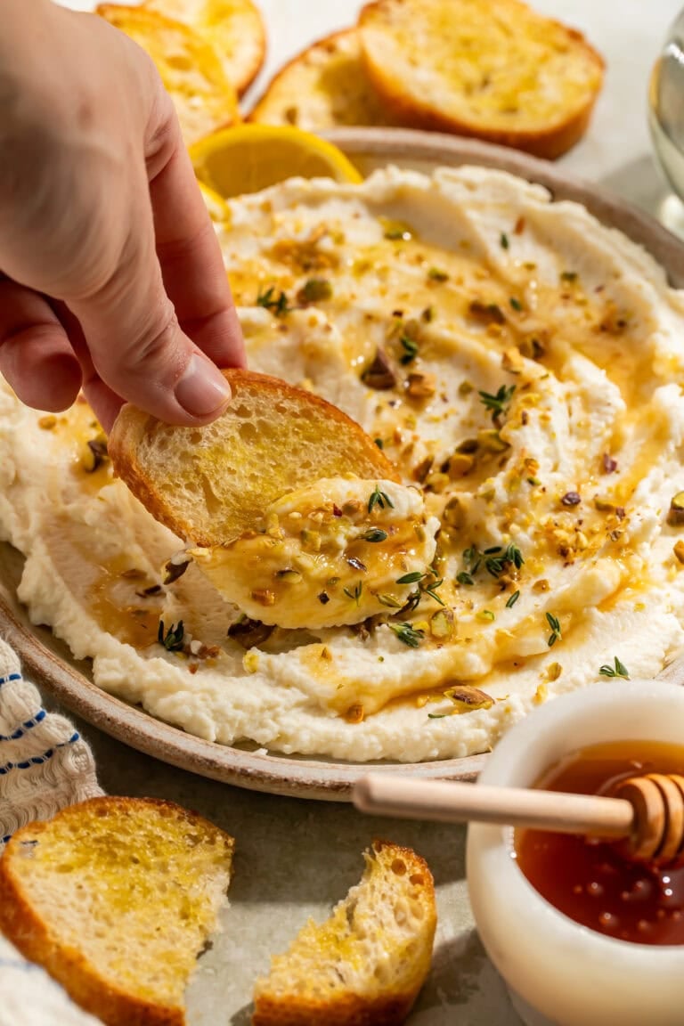 Overhead view of whipped ricotta dip in a shallow bowl, drizzled with honey and topped with chopped pistachios, fresh thyme, and red pepper flakes, with toasted baguette slices and lemon wedges alongside
