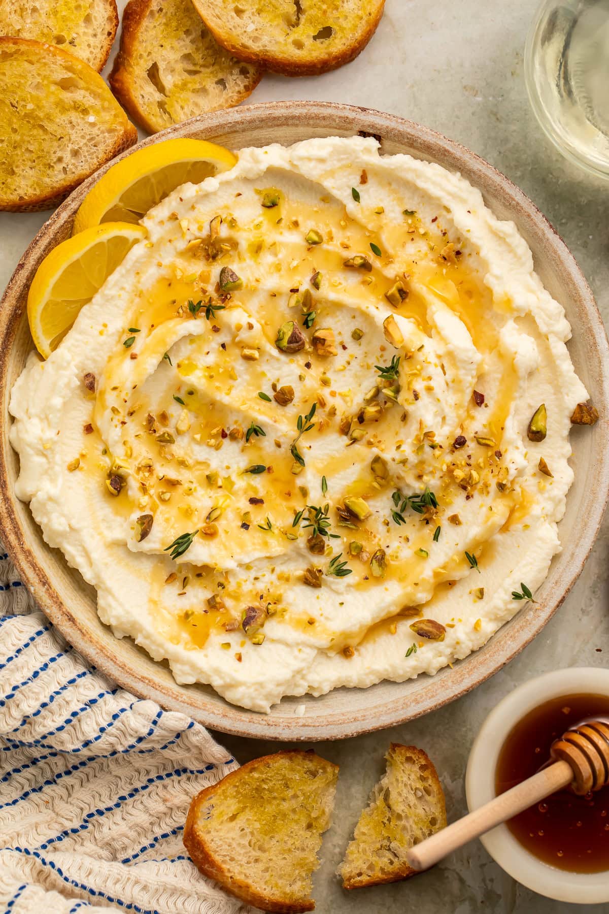 Hand dipping a toasted baguette slice into a shallow bowl of whipped ricotta topped with honey, pistachios, and fresh thyme