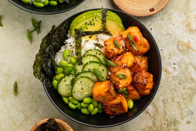Bang bang salmon bowl with crispy salmon bites, white rice, avocado, cucumber, edamame, seaweed, sesame seeds, and green onions in a black bowl