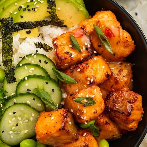 Close-up of bang bang salmon bites served with cucumber, edamame, avocado, and rice in a black bowl