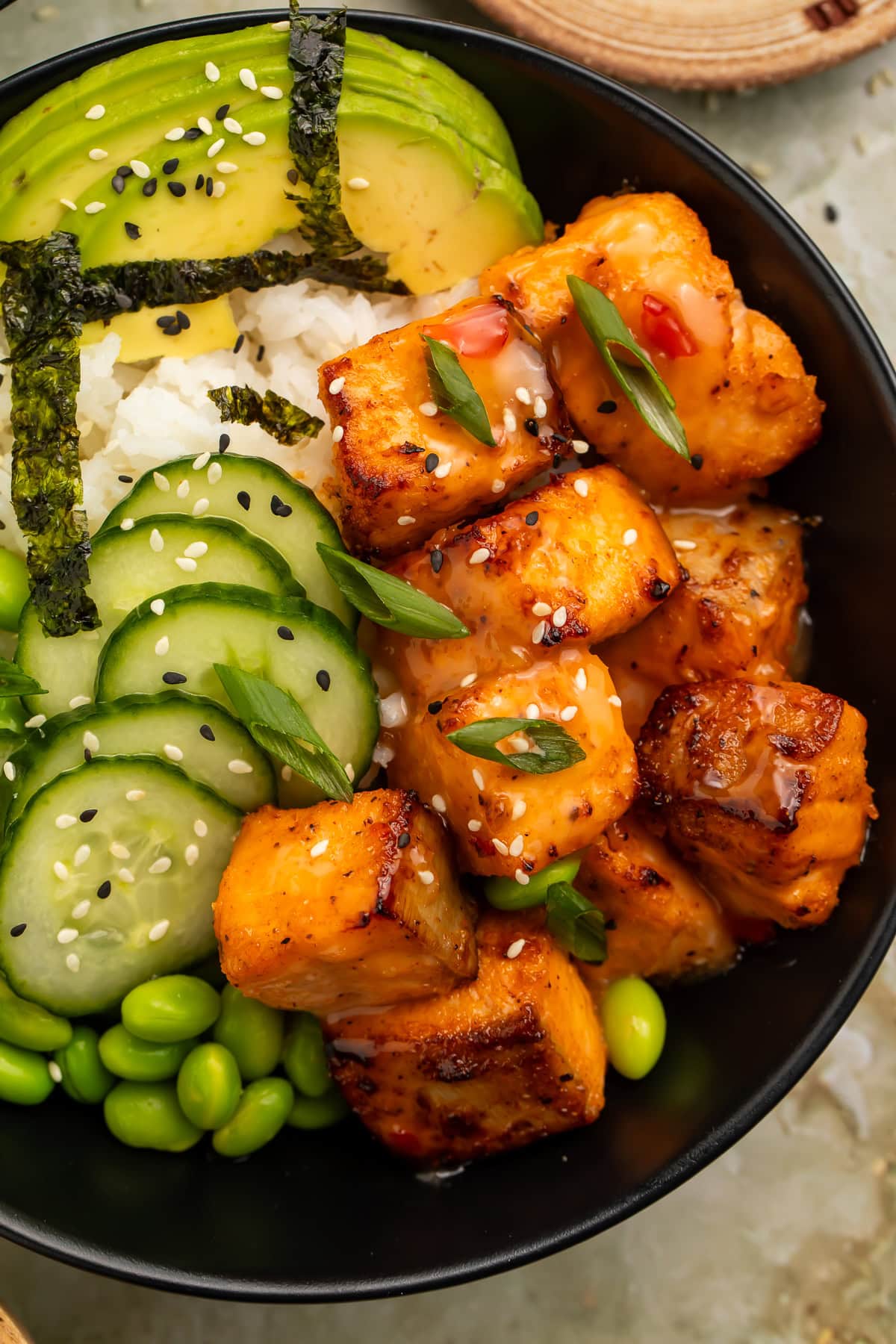 Close-up of bang bang salmon bites served with cucumber, edamame, avocado, and rice in a black bowl