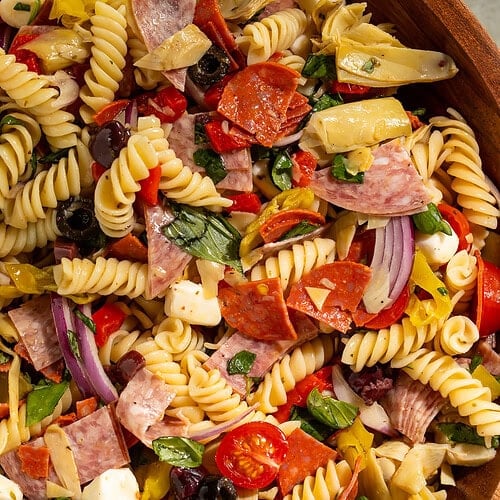 Close up of an antipasto pasta salad in a wooden serving bowl with mozzarella, tomatoes, onions, salami and cured artichokes.