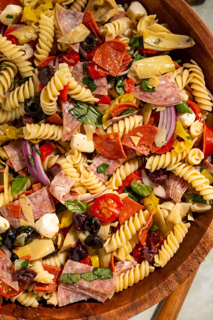 Close up of an antipasto pasta salad in a wooden serving bowl with mozzarella, tomatoes, onions, salami and cured artichokes.