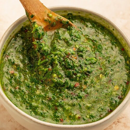 White bowl of chimichurri with a wooden spoon lifting the sauce out of the bowl.