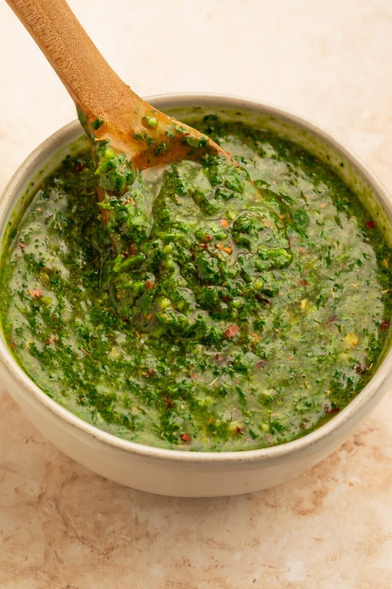 White bowl of chimichurri with a wooden spoon lifting the sauce out of the bowl.