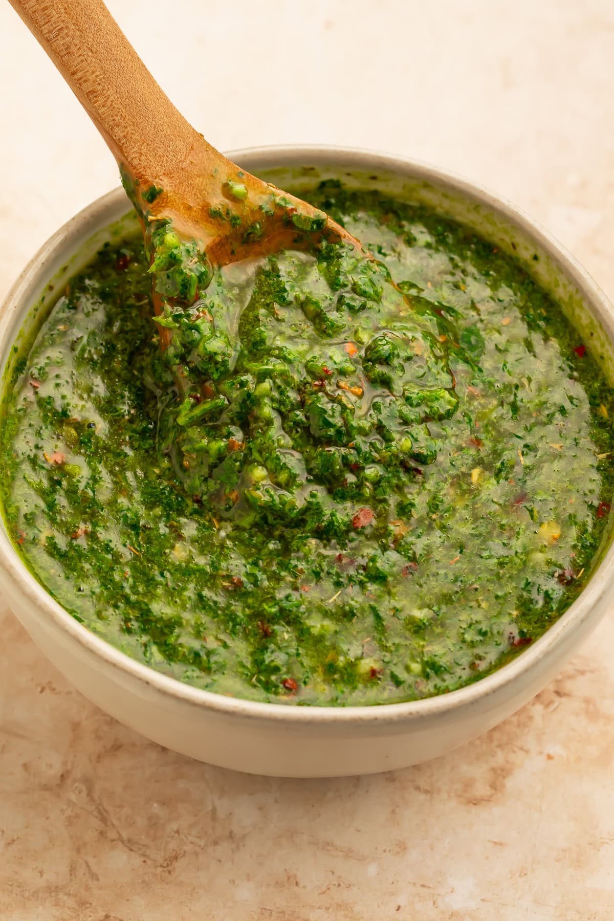 White bowl of chimichurri with a wooden spoon lifting the sauce out of the bowl.