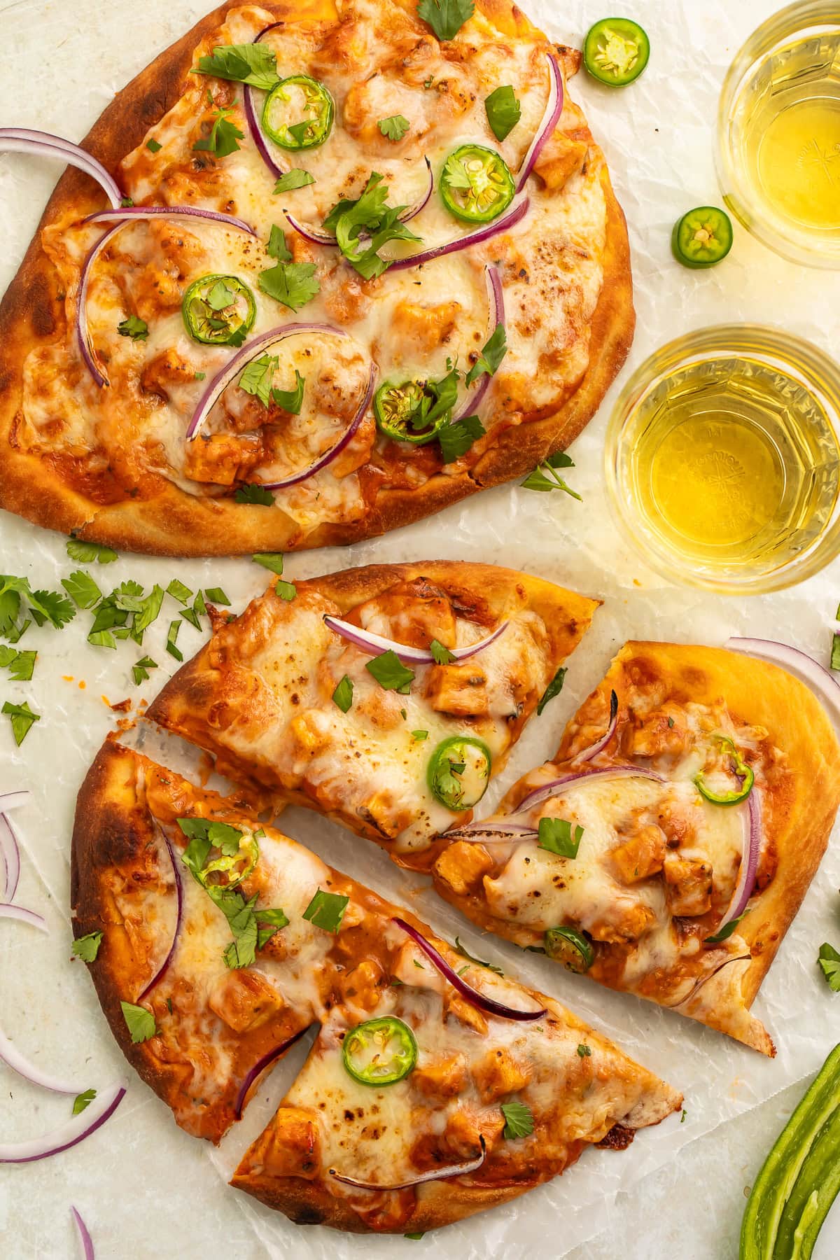 Two naan pizzas topped with melted cheese and chicken tikka masala, scattered with sliced onions, jalapeños and chopped cilantro.