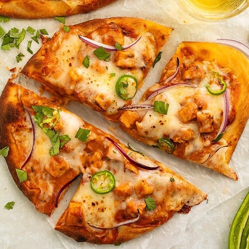 Two chicken tikka masala pizzas on a countertop. The closest one is cut into four, and there is a glass of beer on one side.
