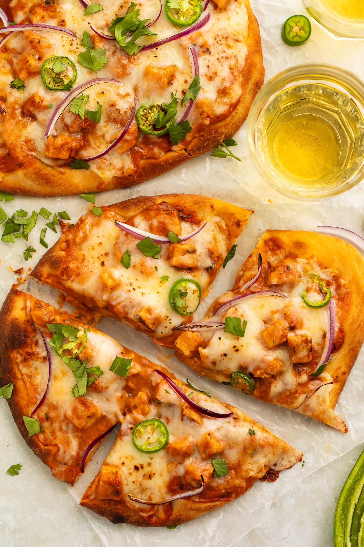 Two chicken tikka masala pizzas on a countertop. The closest one is cut into four, and there is a glass of beer on one side.