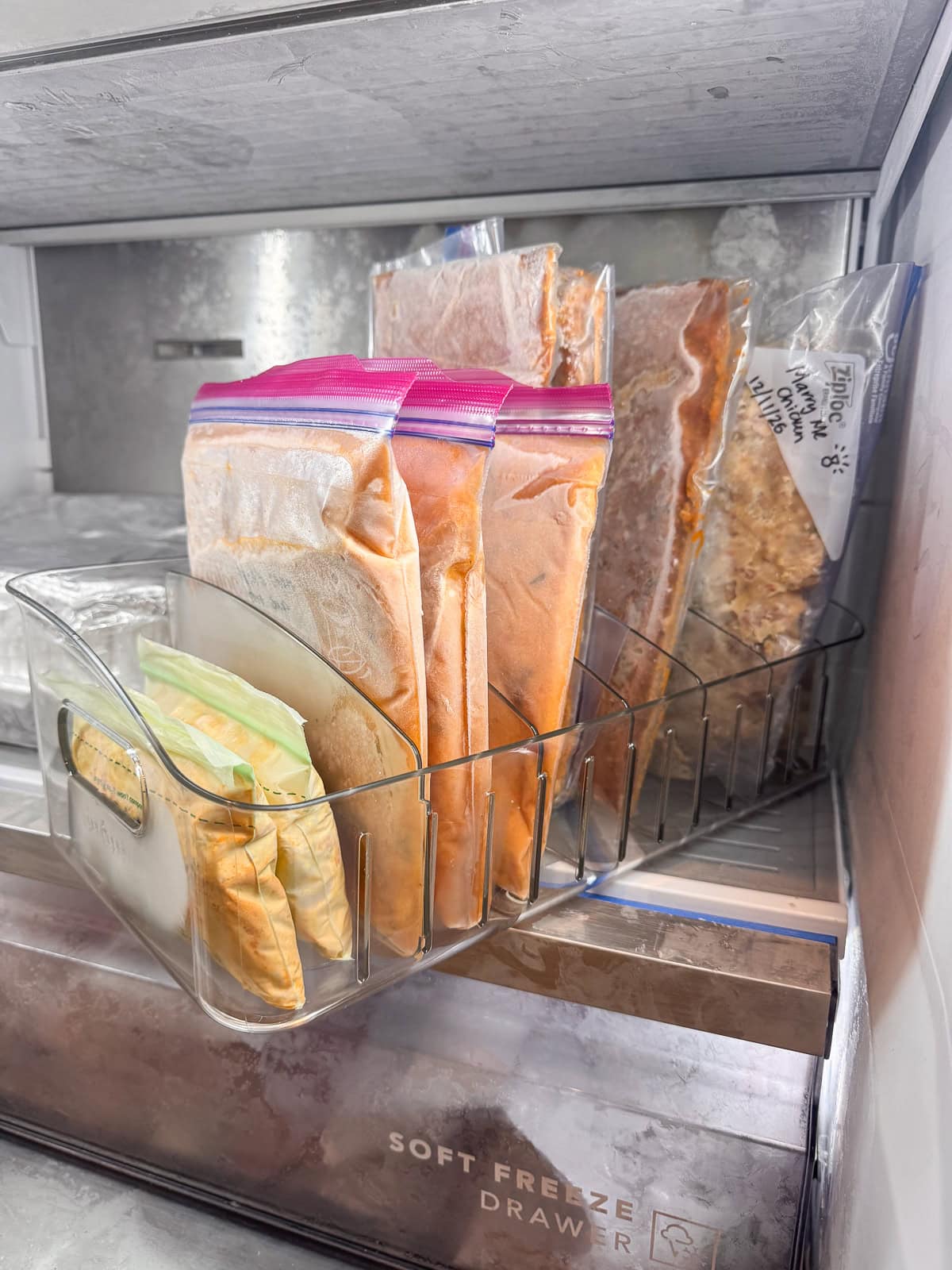 Clear freezer organization bin holding flat frozen meals and sauces in labeled freezer bags inside a freezer drawer.
