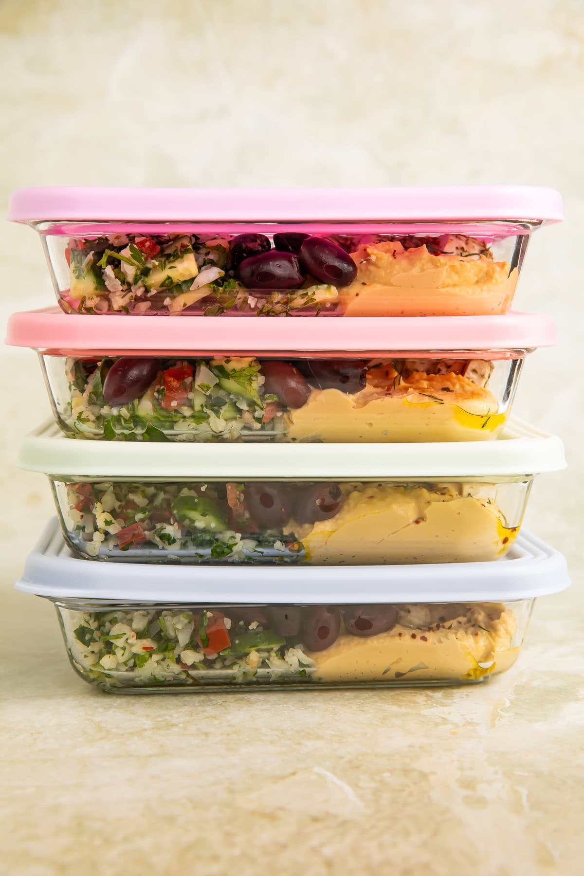Stack of glass meal prep containers with pastel lids filled with Mediterranean-style salads, olives, hummus, and vegetables