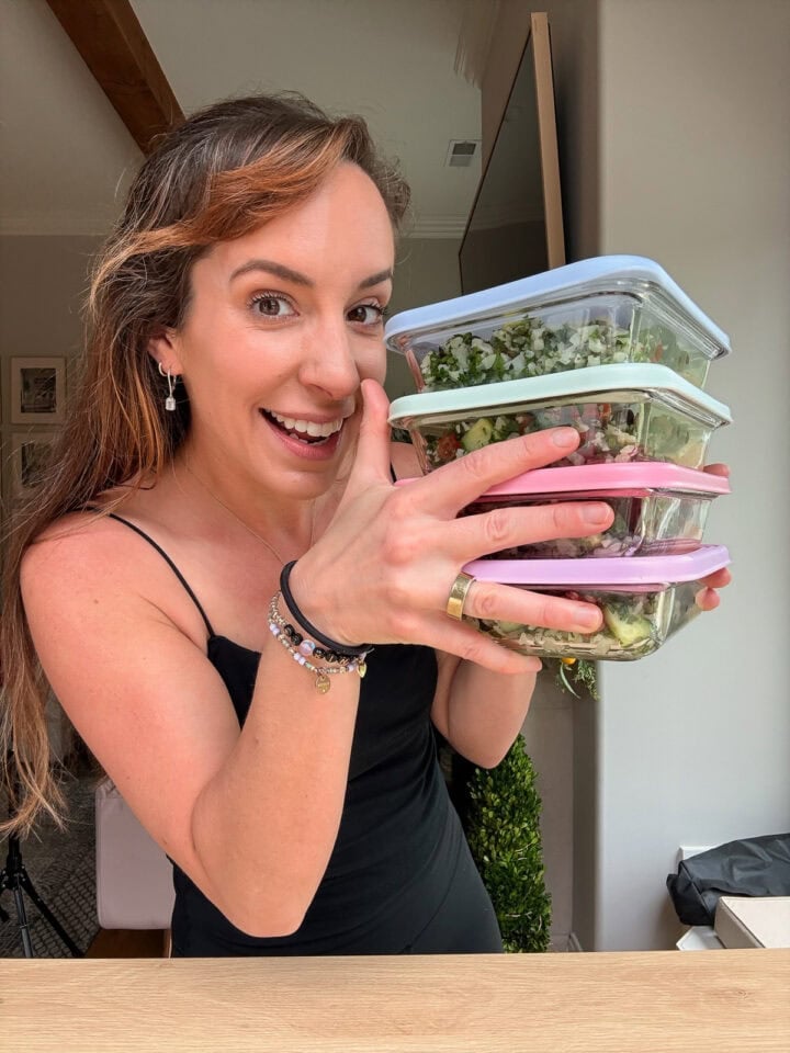 Woman holding pastel-lidded glass meal prep containers filled with Mediterranean-style meals.