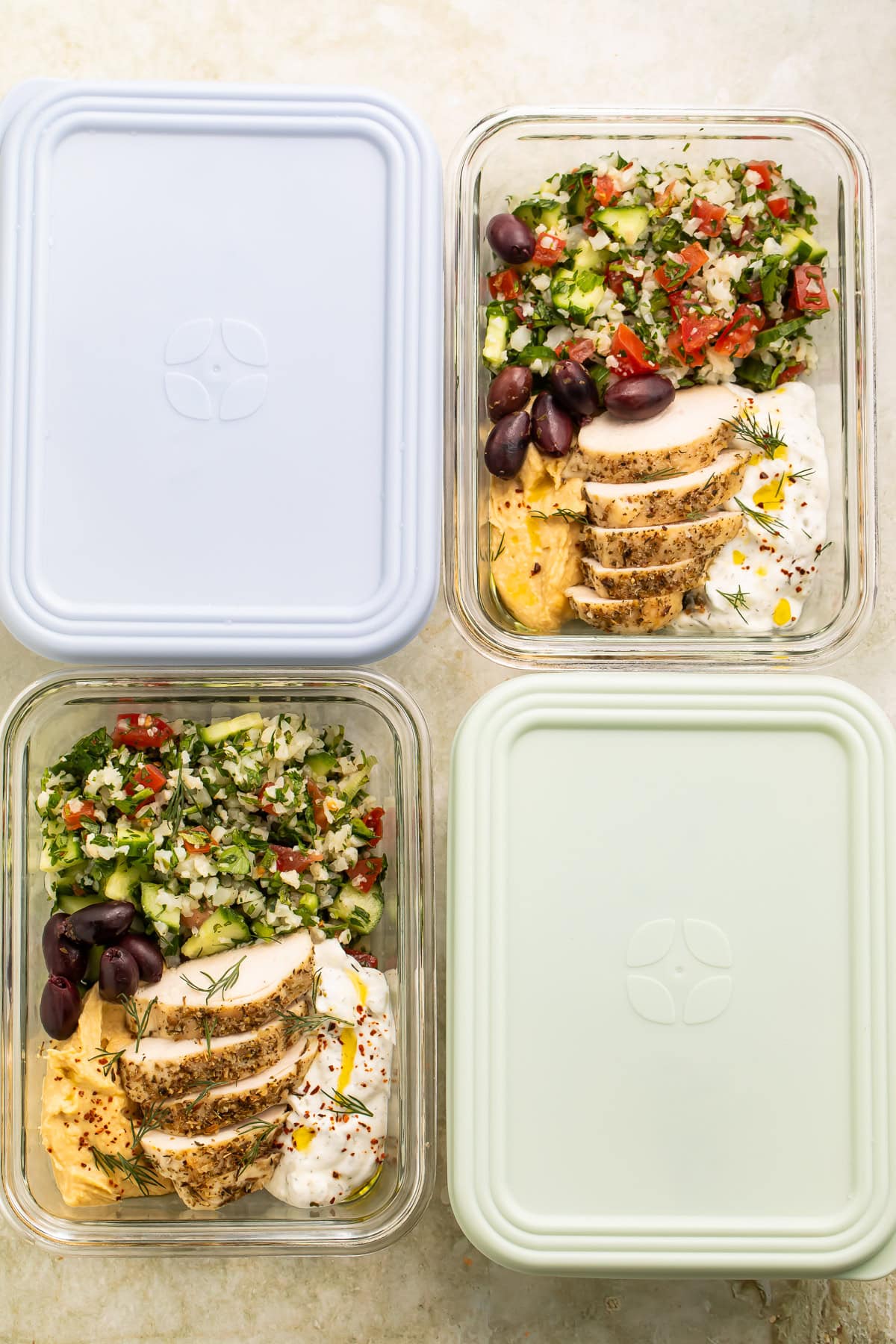 Glass meal prep containers filled with sliced chicken, cauliflower tabbouleh, olives, hummus, and yogurt sauce