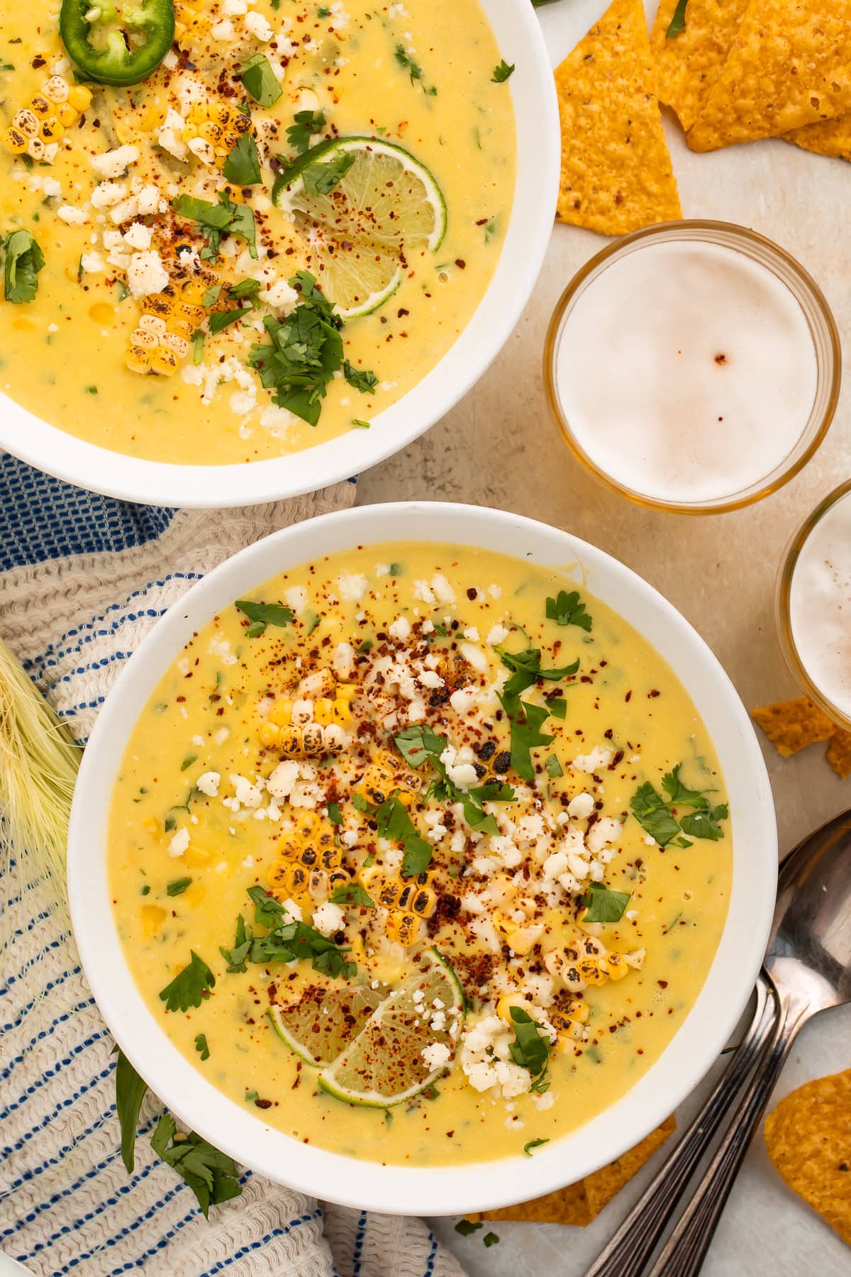 Two bowls of creamy corn soup with street-corn inspired toppings. Tortilla chips and glasses of beer are visible off to one side.