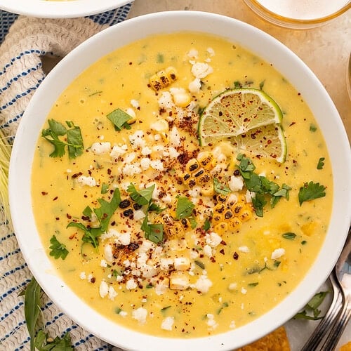 White bowls of creamy yellow Mexican corn soup topped with chili powder, lime slices, cojita and cilantro.