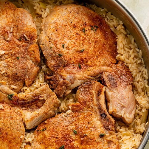 Bone-in pork chops cooked over seasoned rice in a large stainless steel skillet with fresh thyme garnish