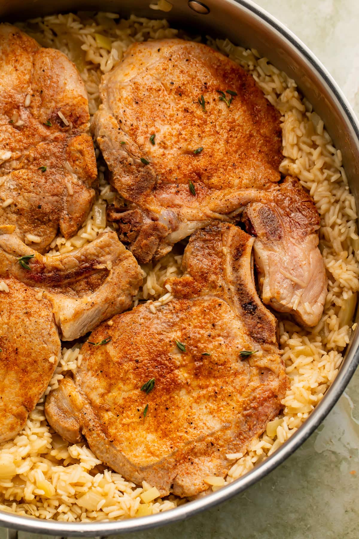 Bone-in pork chops cooked over seasoned rice in a large stainless steel skillet with fresh thyme garnish