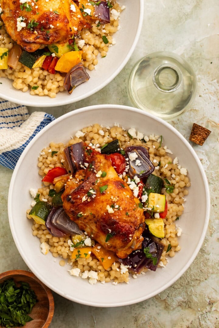 Two bowls of vegetable couscous topped with roasted harissa chicken thighs.