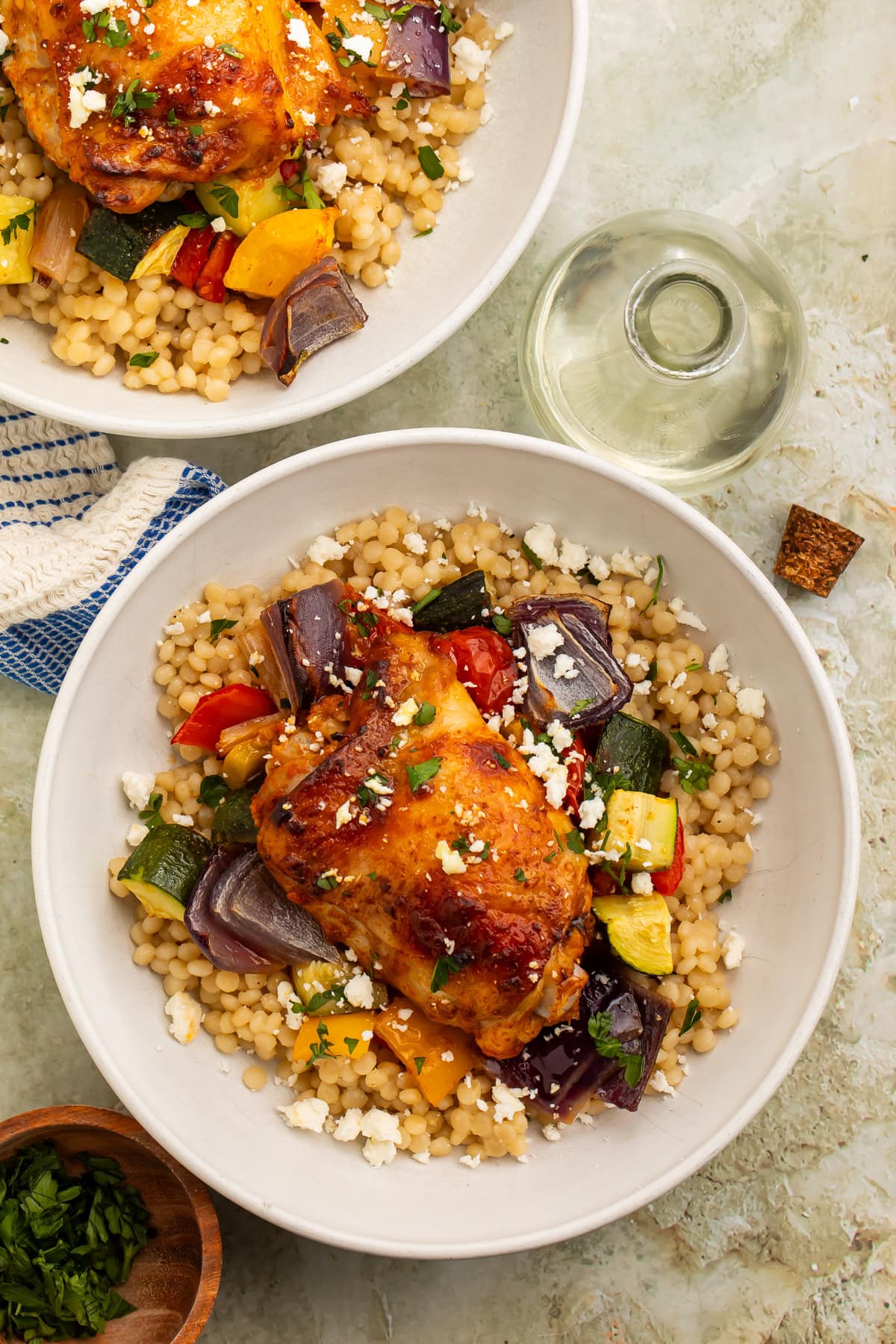 Two bowls of vegetable couscous topped with roasted harissa chicken thighs.