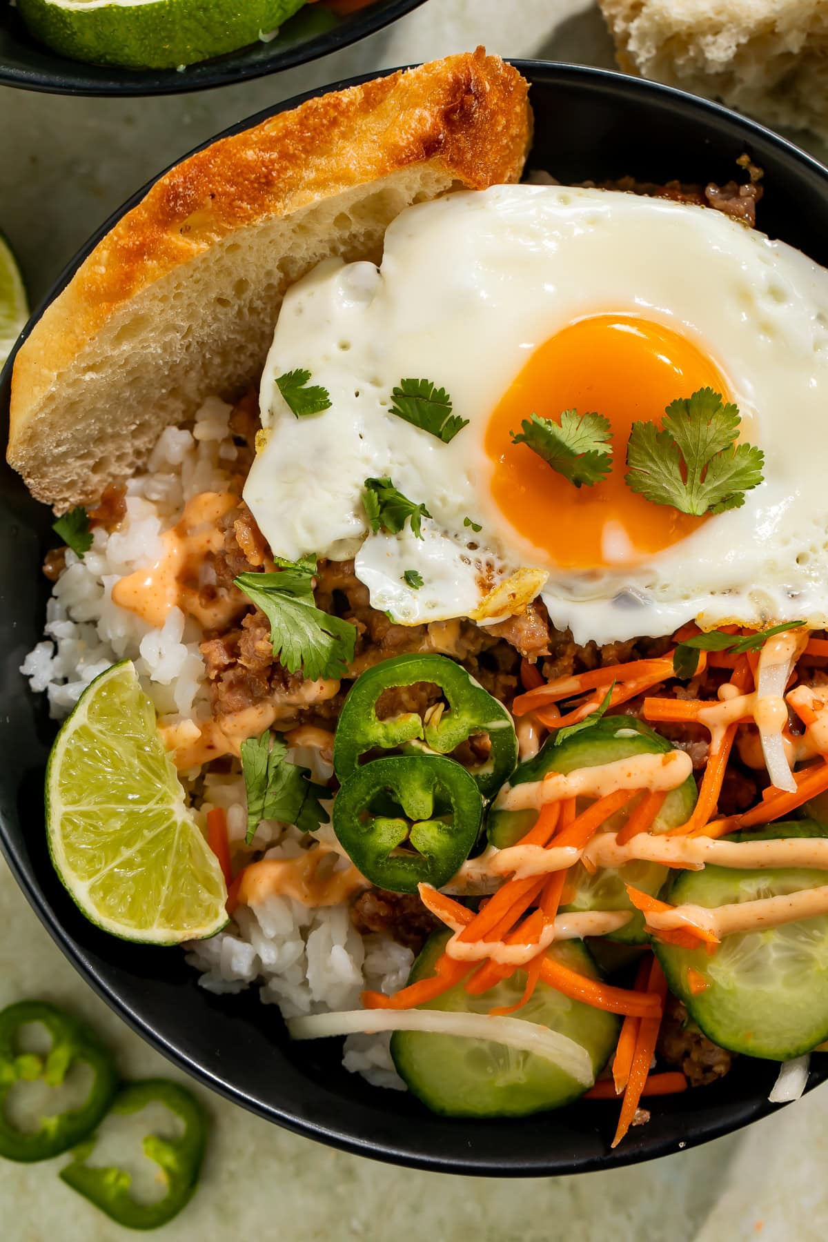 Close-up of a banh mi rice bowl with fried egg, ground meat, white rice, cucumbers, carrots, jalapeños, cilantro, lime wedge, and spicy sauce