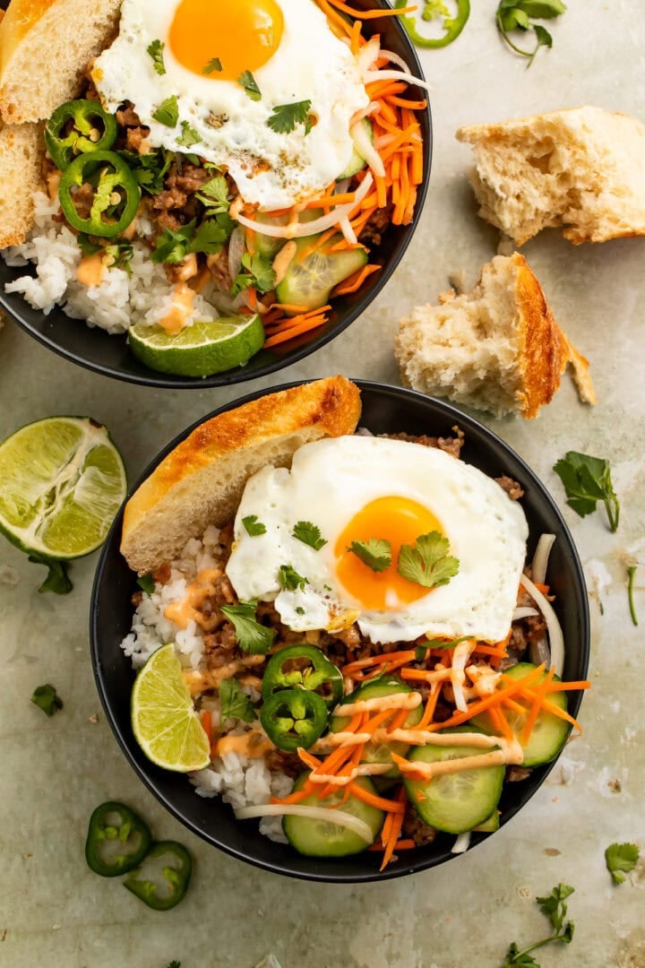 Two banh mi rice bowls topped with fried eggs, ground meat, white rice, cucumbers, shredded carrots, jalapeños, cilantro, lime wedges, and spicy sauce in black bowls with toasted bread on the side.