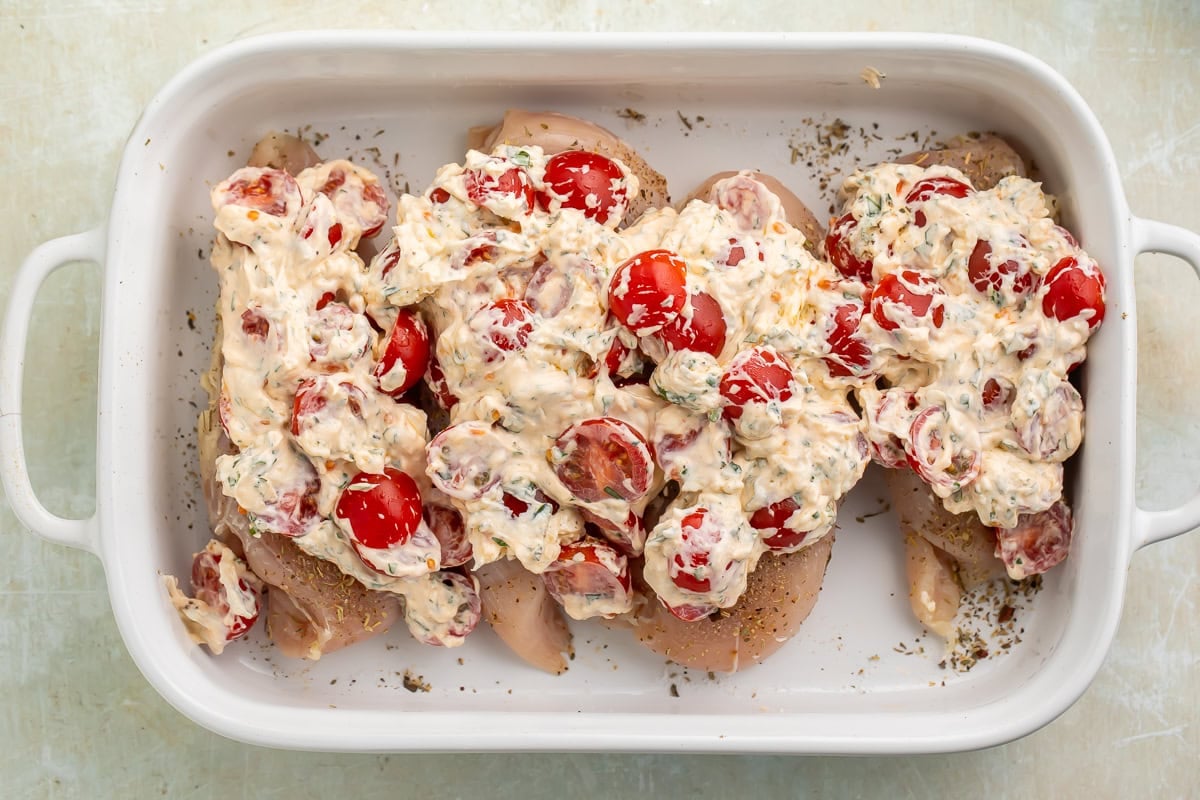 Raw chicken breasts in a white baking dish topped with an uncooked halved cherry tomato creamy topping.